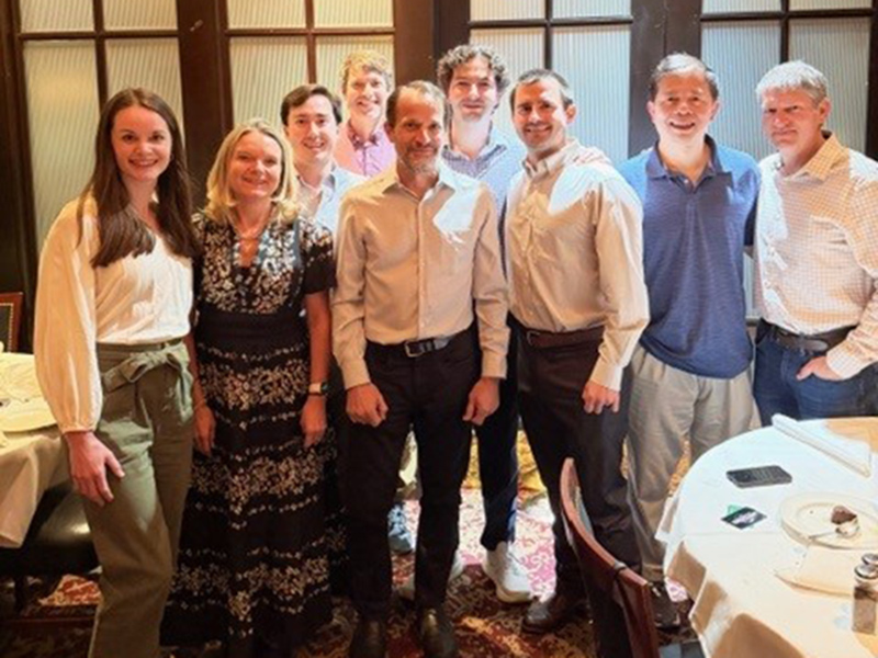 Nine people stand together in a restaurant, smiling at the camera. They are in front of wooden doors with frosted glass; round tables with white cloths are nearby.